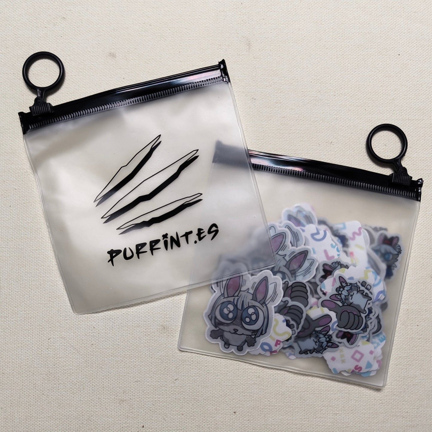 A clear plastic zippered bag with the text 'PURRINT.ES' and logo on it, displayed next to a collection of various stickers and small items inside.